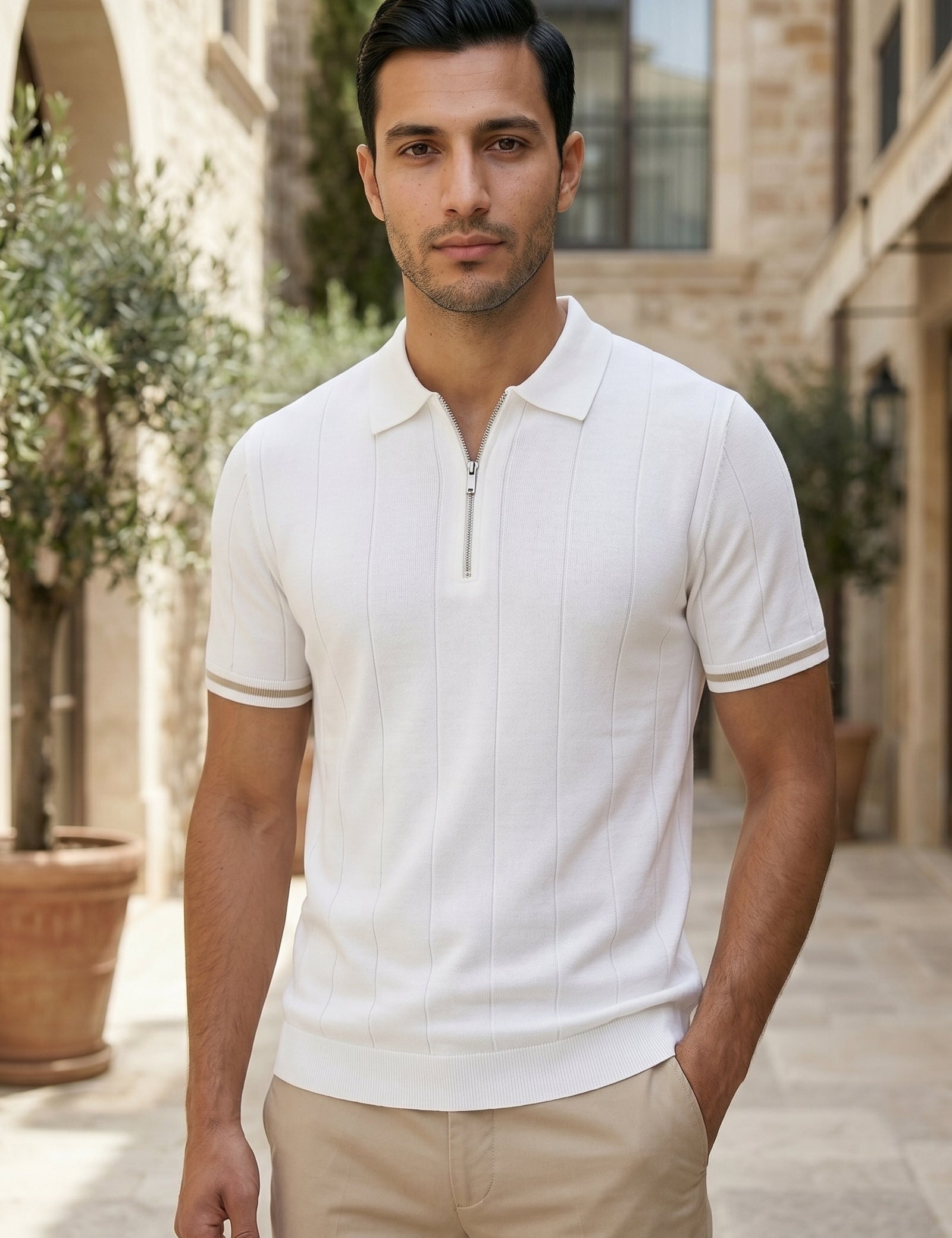 Man wearing a white polo shirt and beige pants standing in an outdoor setting with stone buildings and potted plants.