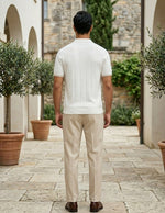 Man wearing a white polo shirt and beige pants standing in an outdoor setting with potted plants and stone walls.