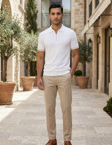 Man wearing a white polo shirt and beige pants standing in an outdoor setting with stone buildings and potted plants.