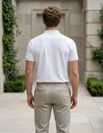 Man wearing a white polo shirt and beige pants standing in front of a building with greenery.
