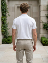 Man wearing a white polo shirt and beige pants standing in front of a building with greenery.