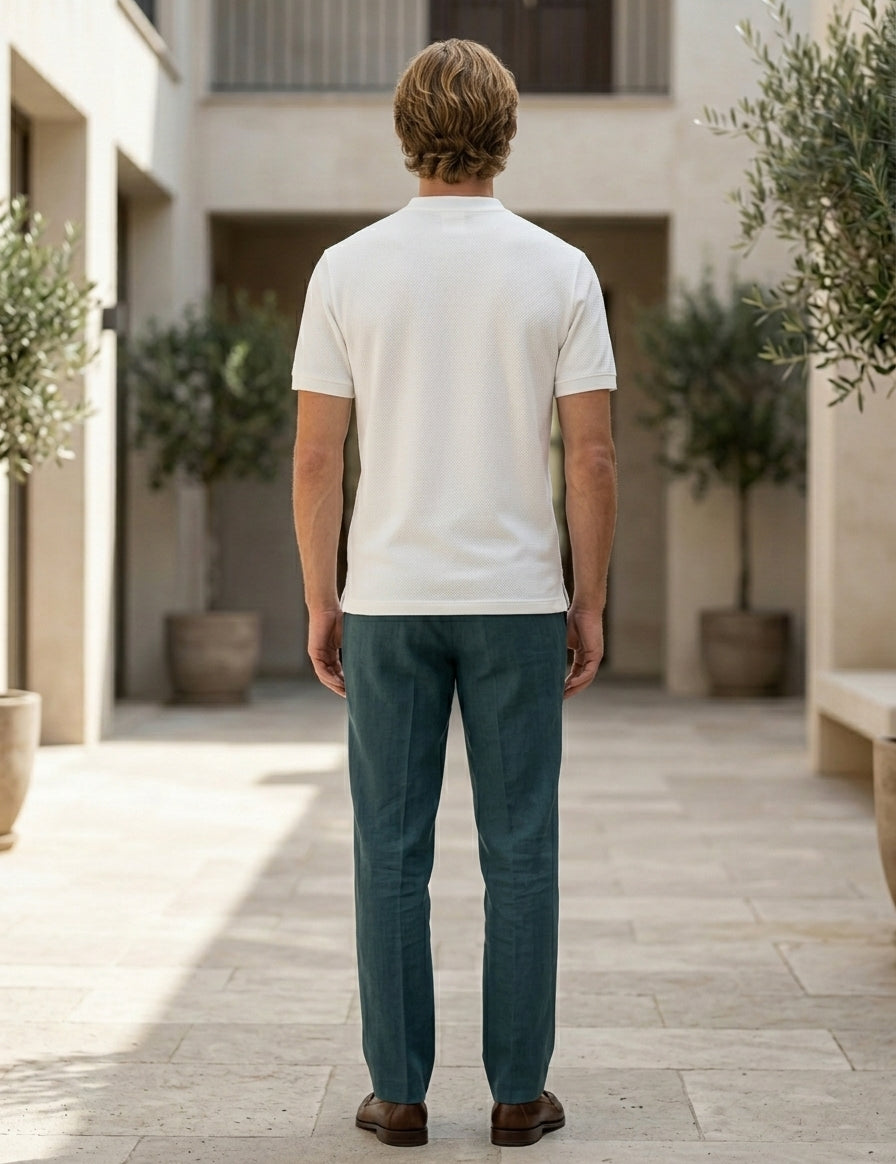 Man wearing a white polo shirt and teal pants standing in an outdoor setting with plants and stone pavement.