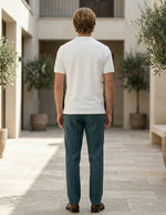 Man wearing a white polo shirt and teal pants standing in an outdoor setting with plants and stone pavement.