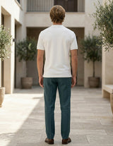 Man wearing a white polo shirt and teal pants standing in an outdoor setting with plants and stone pavement.