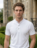 Man wearing a white polo shirt standing in front of a classical building with greenery.