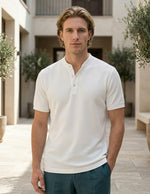 Man wearing a white polo shirt standing outdoors with plants and buildings in the background