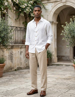 Man wearing a white shirt and beige pants standing in an outdoor setting with plants and stone architecture.