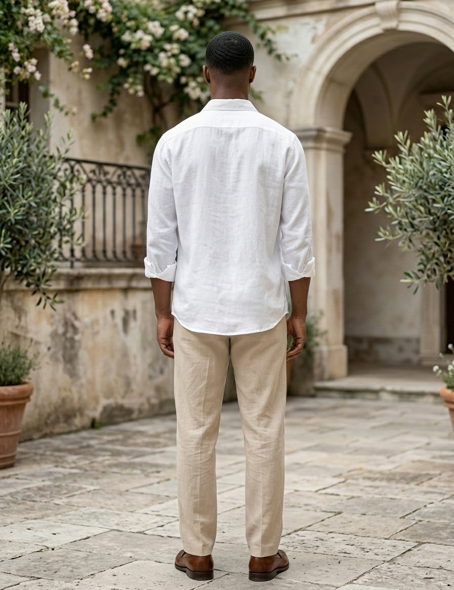 Man wearing a white shirt and beige pants standing in an outdoor setting with stone pavement and plants.