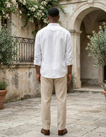 Man wearing a white shirt and beige pants standing in an outdoor setting with stone pavement and plants.