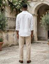 Man wearing a white shirt and beige pants standing in an outdoor setting with stone pavement and plants.
