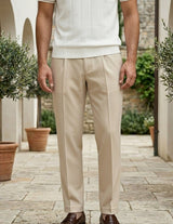 Man wearing a white shirt and beige pants standing outdoors with plants and stone walls in the background.