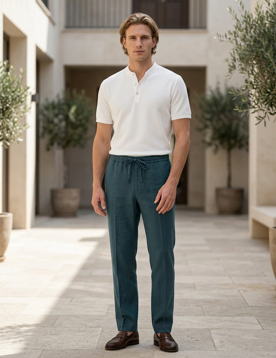Man wearing a white shirt and teal pants standing in an outdoor setting with plants and stone pavement.