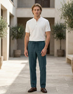 Man wearing a white shirt and teal pants standing in an outdoor setting with plants and stone pavement.