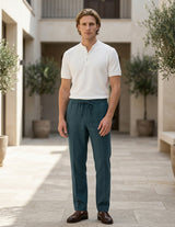 Man wearing a white shirt and teal pants standing in an outdoor setting with plants and stone pavement.