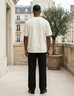 Man wearing a white t-shirt and black pants standing on a stone patio with classical architecture in the background.