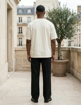 Man wearing a white t-shirt and black pants standing on a stone patio with classical architecture in the background.
