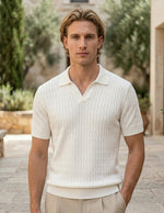 Man wearing a white textured shirt standing outdoors with greenery and stone architecture in the background