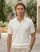 Man wearing a white textured shirt standing outdoors with greenery and stone architecture in the background