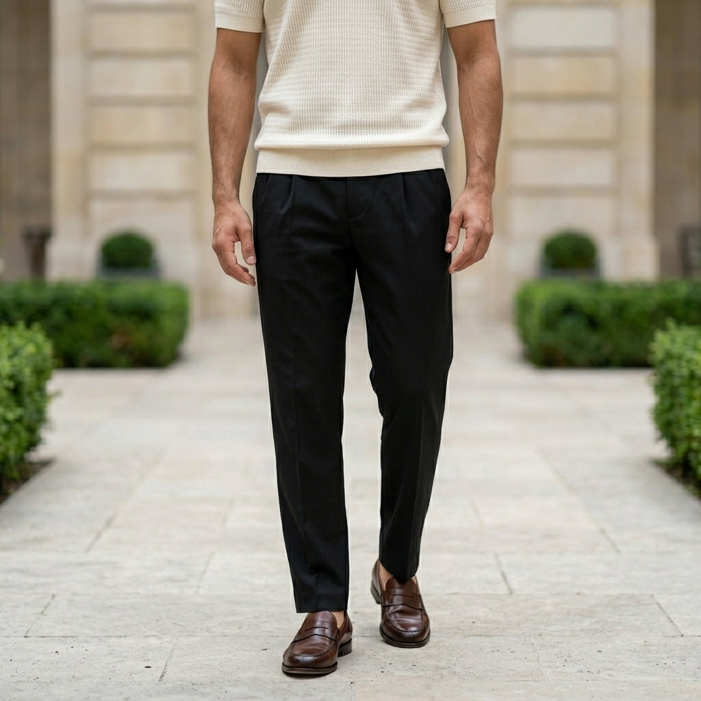 Man wearing black pants and brown shoes standing on a stone path with greenery and a building in the background.