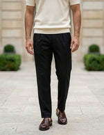 Man wearing black pants and brown shoes standing on a stone path with greenery and a building in the background.