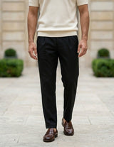Man wearing black pants and brown shoes standing on a stone path with greenery and a building in the background.