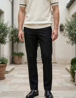Person wearing a beige polo shirt and black pants standing in an outdoor setting with plants.
