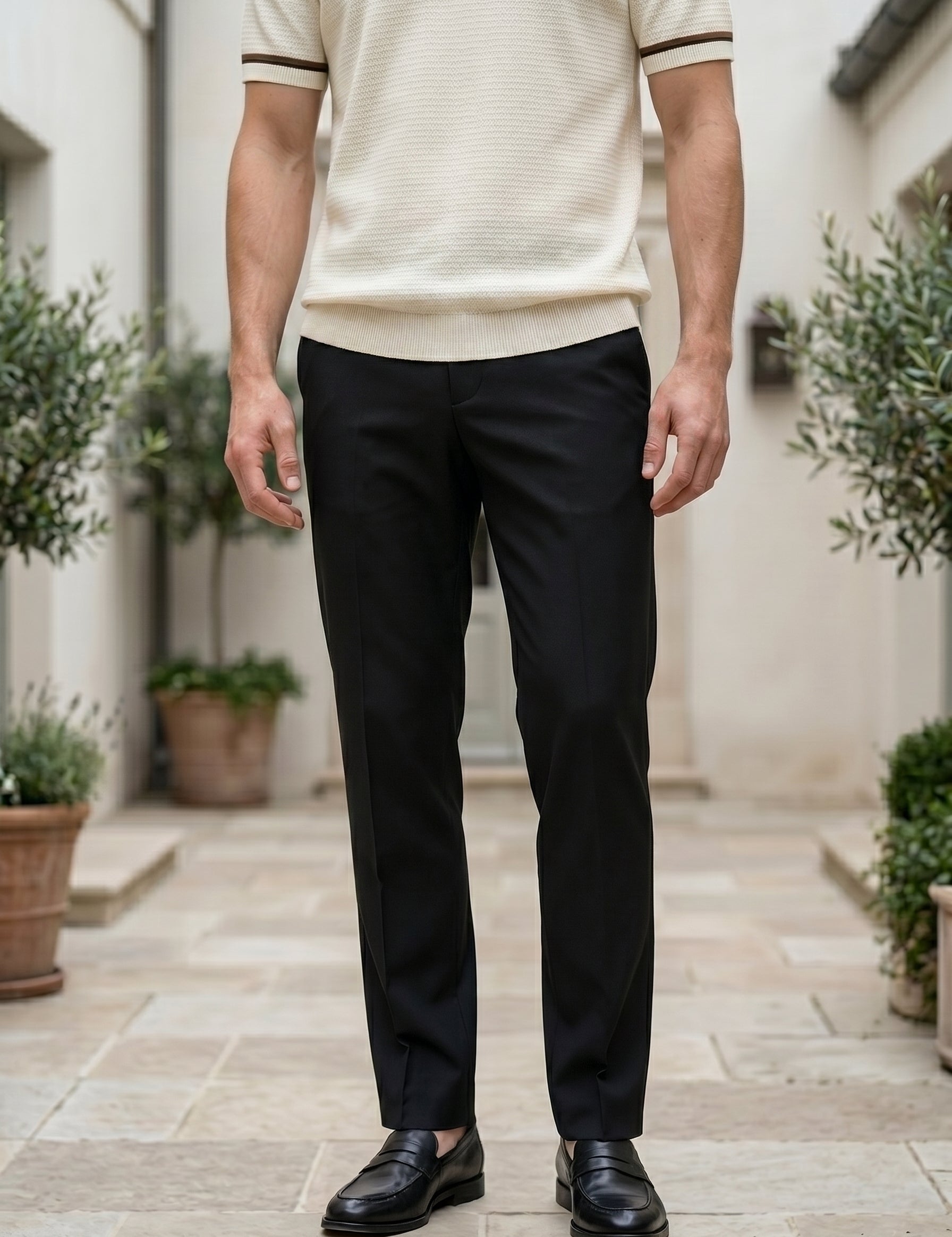 Person wearing a beige polo shirt and black pants standing in an outdoor setting with plants.