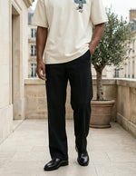 Person wearing a beige shirt and black pants standing on a stone pavement with buildings and a tree in the background.