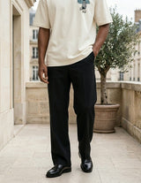Person wearing a beige shirt and black pants standing on a stone pavement with buildings and a tree in the background.
