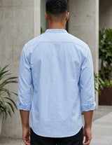 Person wearing a light blue shirt standing in an outdoor setting with arches and plants.