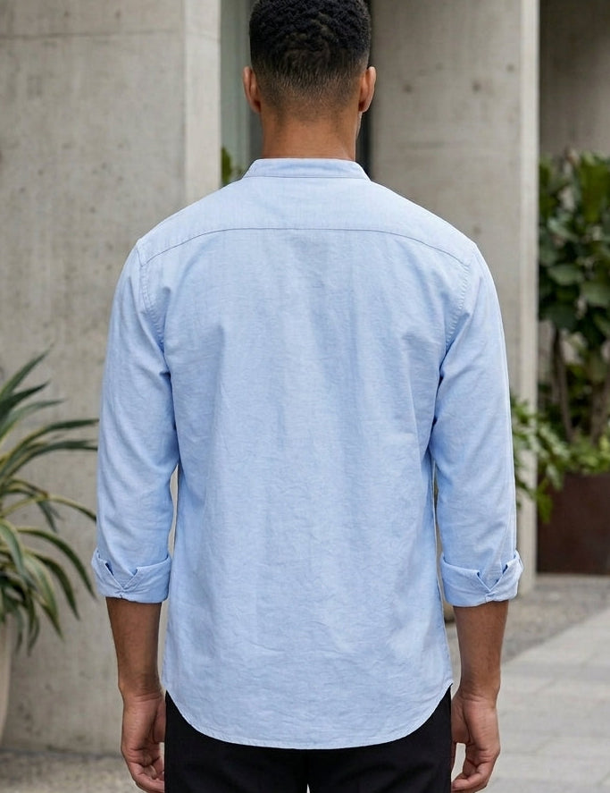 Person wearing a light blue shirt standing in an outdoor setting with arches and plants.