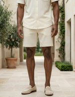 Person wearing a white outfit standing in an outdoor setting with plants and stone pavement.