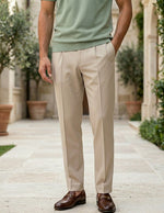 Person wearing beige pants and a green shirt standing outdoors with plants and stone pavement in the background.