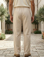 Person wearing beige pants and a light-colored shirt standing on a stone pavement with plants in the background.