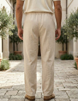 Person wearing beige pants and a light-colored shirt standing on a stone pavement with plants in the background.