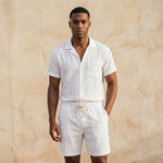 Man wearing a white linen shirt and shorts against a beige wall