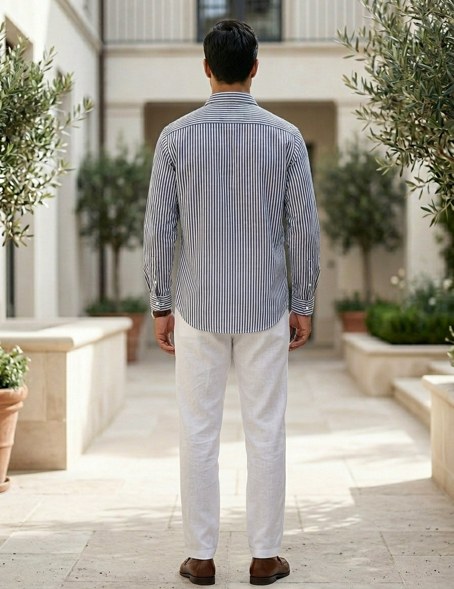 Man wearing a striped shirt and white pants standing in an outdoor setting with plants.