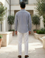 Man wearing a striped shirt and white pants standing in an outdoor setting with plants.