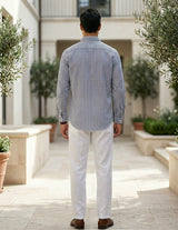 Man wearing a striped shirt and white pants standing in an outdoor setting with plants.