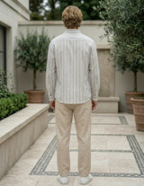 Man wearing a striped shirt and beige pants standing in an outdoor setting with plants and a building.