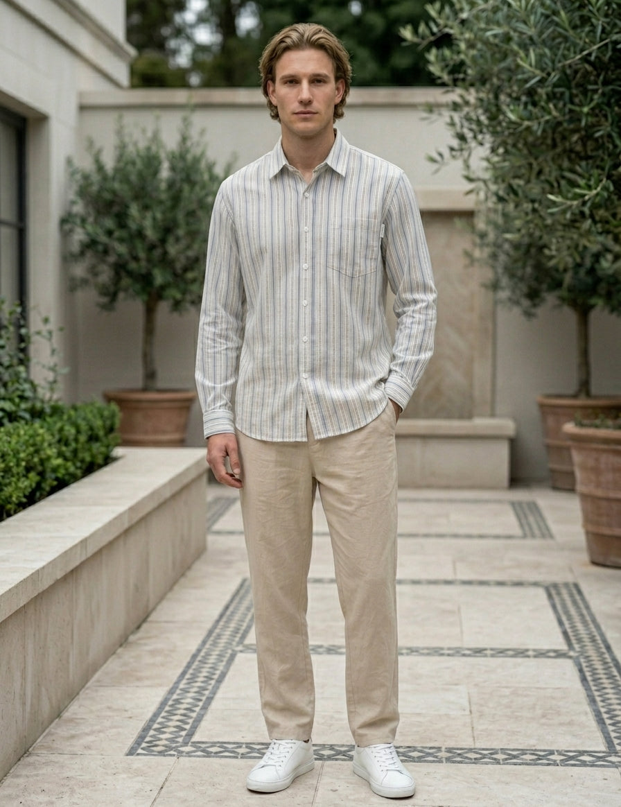 Man wearing a striped shirt and beige pants standing in an outdoor setting with plants and a building.