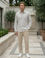 Man wearing a striped shirt and beige pants standing in an outdoor setting with plants and a building.