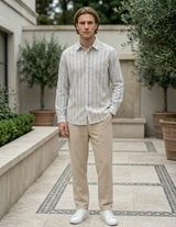 Man wearing a striped shirt and beige pants standing in an outdoor setting with plants and a building.