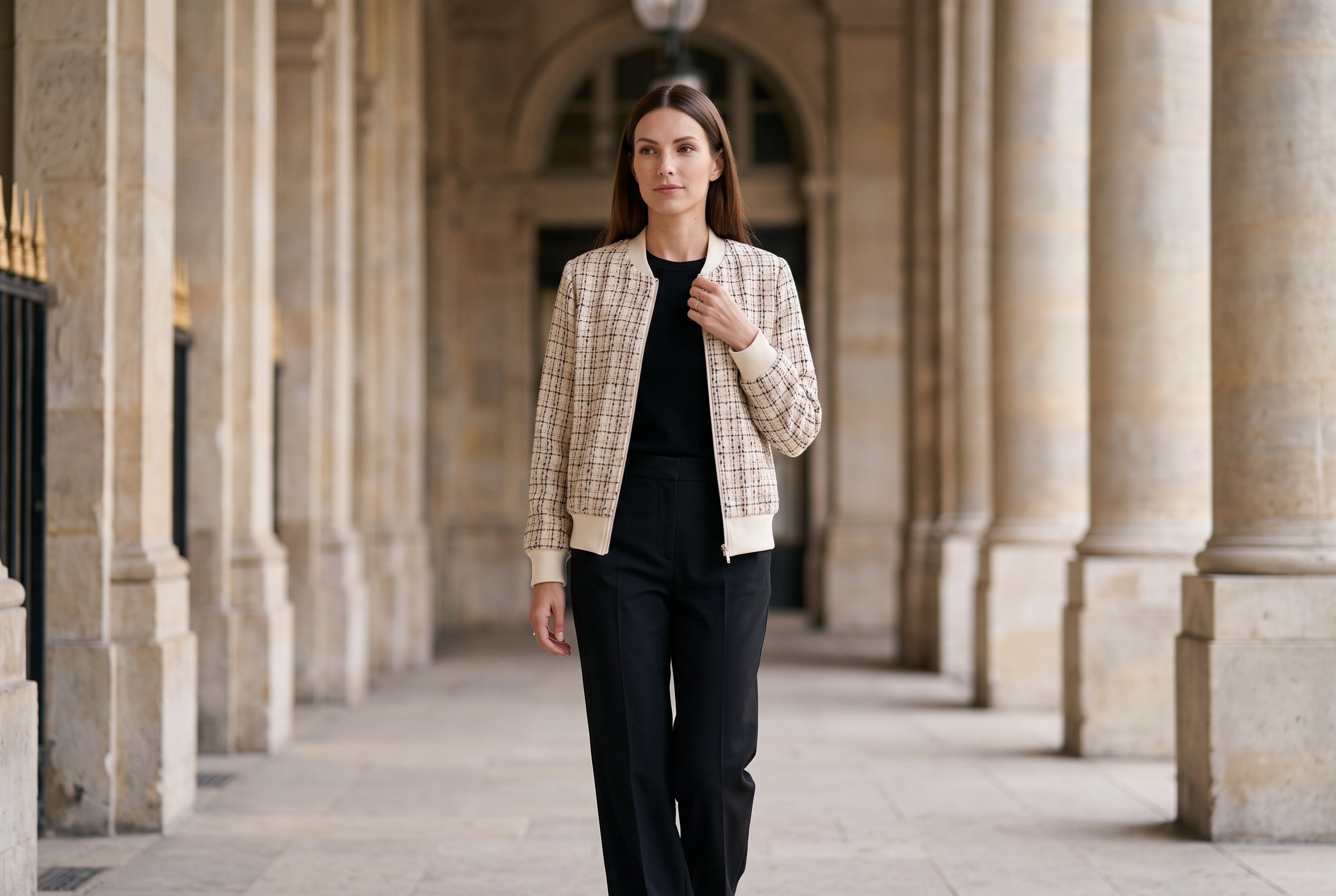 Woman in a cream tweed bomber jacket walking under the stone arcades of a Paris garden palace