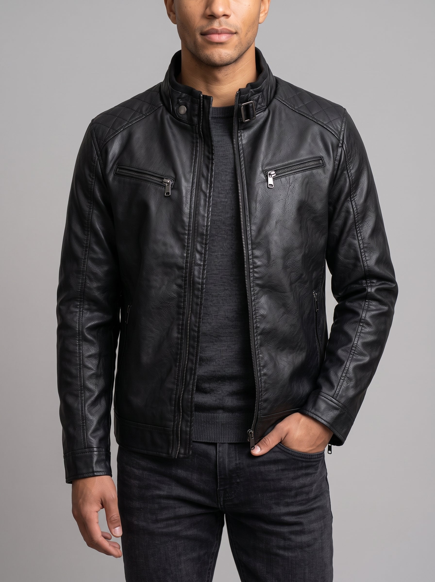 A man wearing a black leather jacket over a dark gray shirt and dark jeans, standing against a solid gray studio background.
