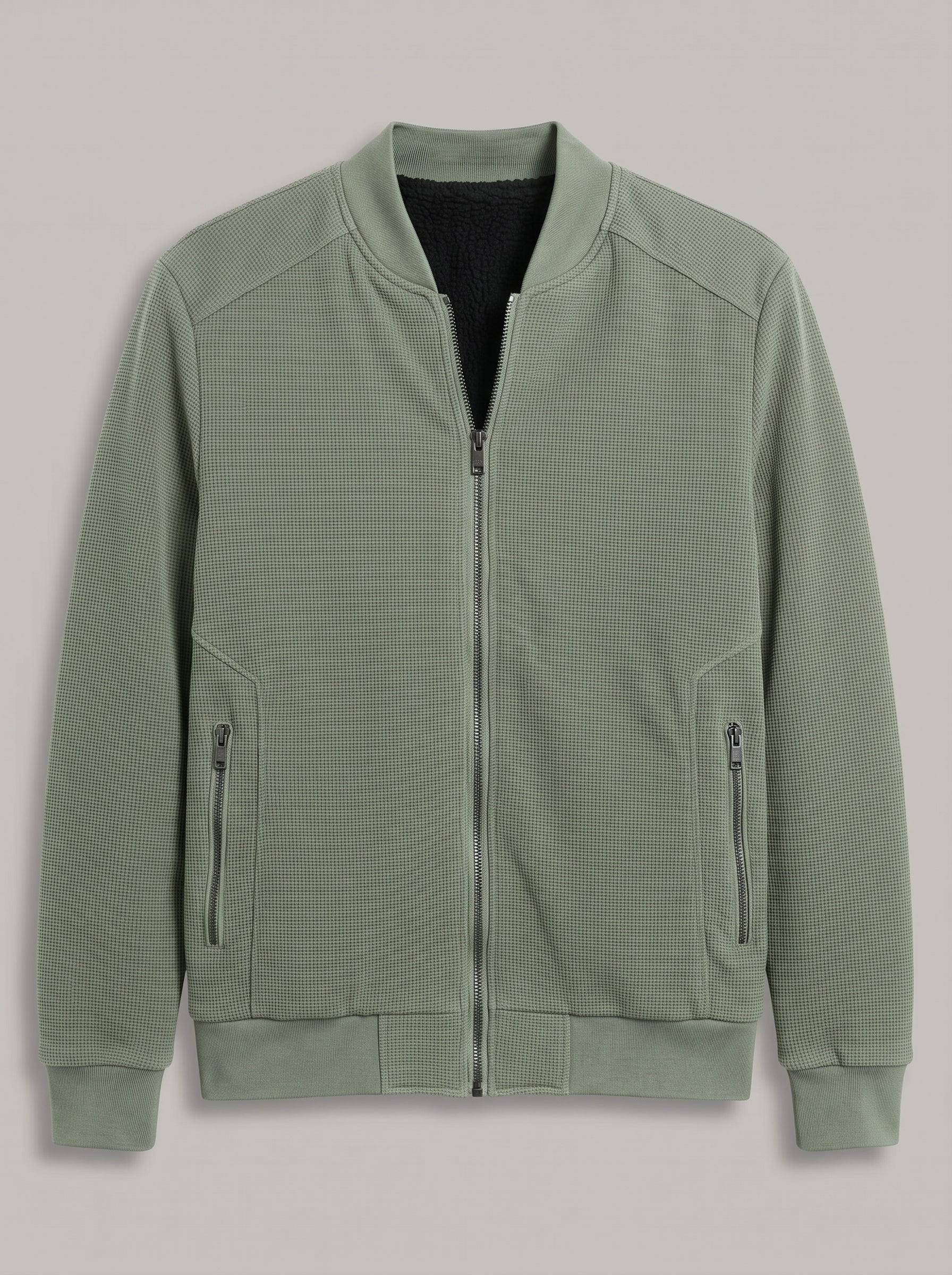 A sage green zip-up bomber jacket with a textured waffle knit fabric, ribbed cuffs and hem, and zippered hand pockets, shown against a neutral gray background.