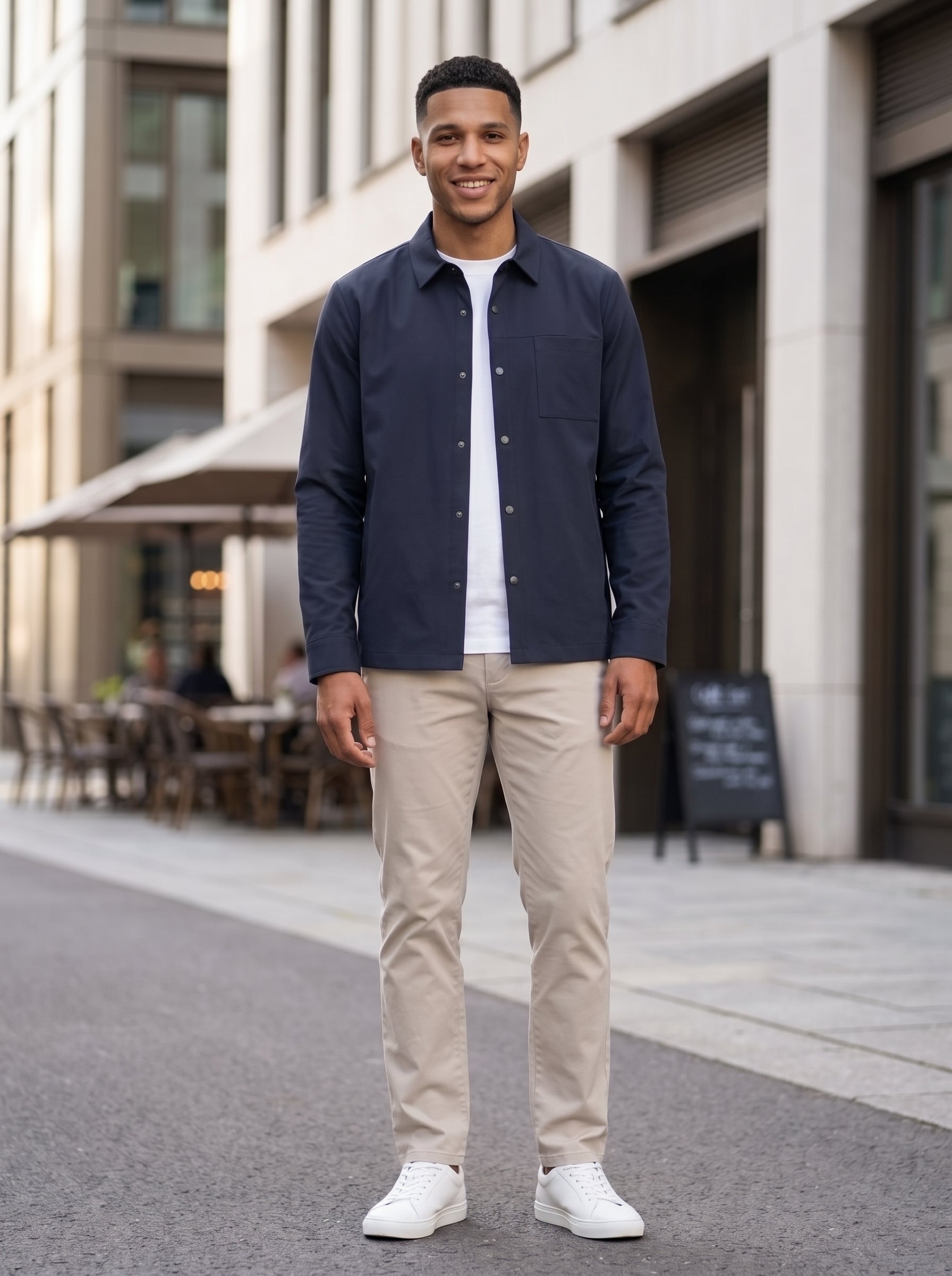 A man standing outdoors on a city street wearing a navy blue overshirt over a white t-shirt, beige trousers, and white sneakers.