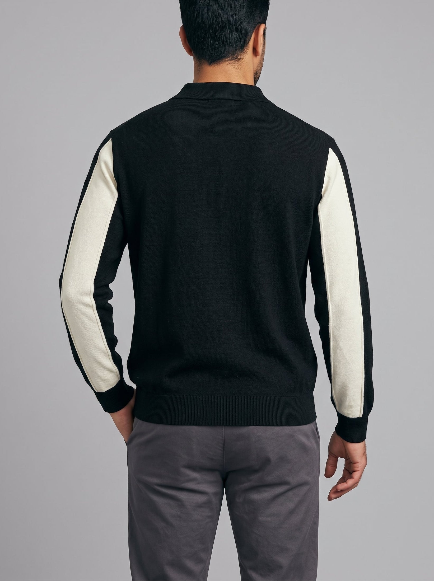 A man viewed from the back wearing a black knit sweater with contrasting off-white sleeves, paired with gray trousers, against a solid gray studio background.