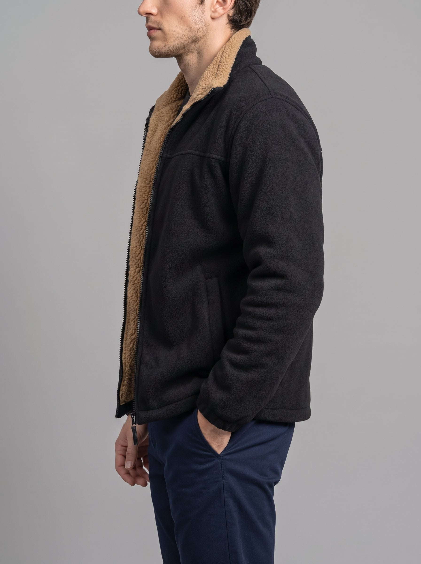 A man shown in profile wearing a black fleece jacket with a tan sherpa lining and navy blue pants, photographed against a solid gray studio background.