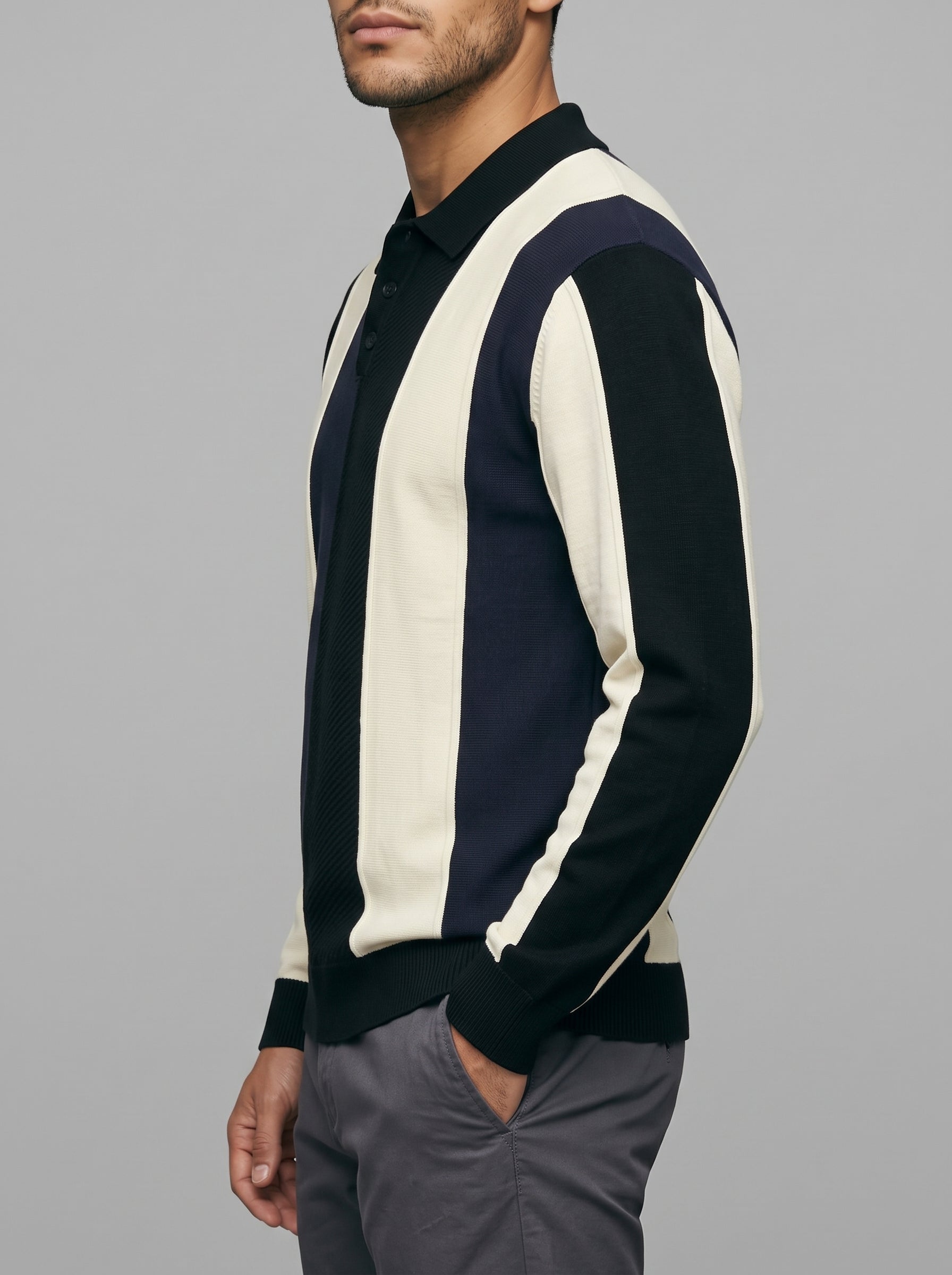 A man wearing a color-block knit polo shirt with vertical stripes in cream, navy, and black, paired with gray trousers, shown from the side against a solid gray background.
